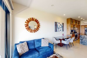A modern living room with a blue sofa, a dining table with chairs, a kitchen area, and a large mirror on the wall. - Ground-floor condo with ocean view, pool, gym, hot tub, & washer/dryer (Cabo San Lucas)