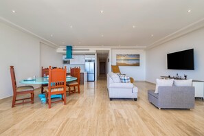 A modern living room with a dining area, a grey sofa, a flat-screen TV, and a kitchen in the background. - Charming 2 BR Condo | Ocean Views, Pool & Gym (Cabo San Lucas)