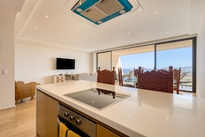 A modern kitchen with a built-in stove, a flat-screen TV, and a large window offering a view of the outdoors. - Charming 2 BR Condo | Ocean Views, Pool & Gym (Cabo San Lucas)