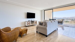 Een moderne woonkamer met een bank, rieten stoelen, een flatscreen televisie, een kast en een groot raam met uitzicht op de stad.