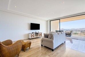 A modern living room with a sofa, wicker chairs, a flat-screen TV, a cabinet, and a large window offering a city view. - Charming 2 BR Condo | Ocean Views, Pool & Gym (Cabo San Lucas)