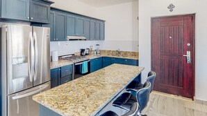 Eine Küche mit dunkelblauen Schränken, einem Edelstahl-Kühlschrank, einer Arbeitsplatte aus Granit und einer dunklen Holztür.