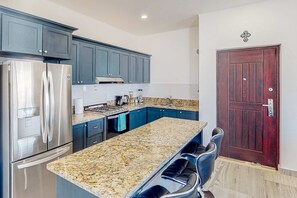 Eine Küche mit dunkelblauen Schränken, einem Edelstahl-Kühlschrank, einer Arbeitsplatte aus Granit und einer dunklen Holztür.