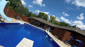 Una piscina con tobogán, una terraza de madera y un edificio de ladrillo al fondo.