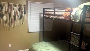 Dormitorio con literas, una colcha verde, un adorno de macramé en la pared y una ventana con persianas.