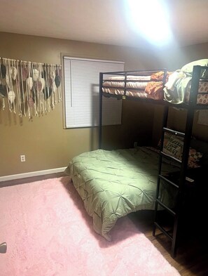 Quarto com beliche, colcha verde, decoração de macramé na parede e janela com persianas.