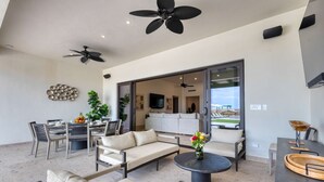 Un'ampia zona giorno con divano, poltrone, tavolo da pranzo e tavolino. Sono presenti ventilatori a soffitto, un'ampia finestra e vista all'esterno.