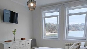 Une chambre à coucher comprenant un lit, une commode, un téléviseur et deux fenêtres donnant sur l’extérieur.
