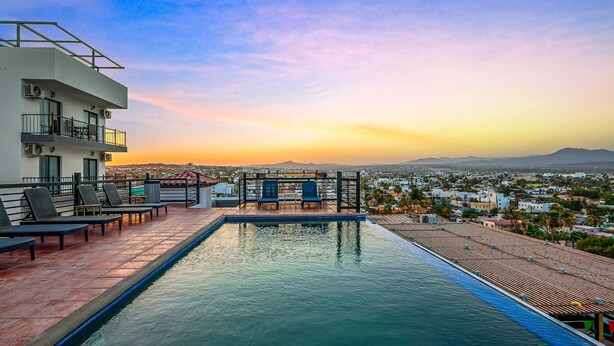 Una piscina en la azotea con sillones, una pérgola y vistas a la ciudad al atardecer.