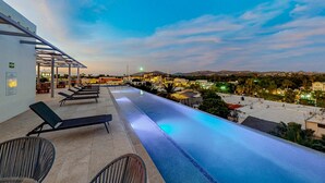 Ein Infinity-Pool mit Liegestühlen, einer überdachten Sitzgelegenheit und Blick auf eine Stadt im Abendrot.