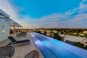 An infinity pool with lounge chairs, a covered seating area, and a view of a town at dusk. - Downtown condo with great views, shared pool, hot tub, & grill (San José del Cabo)