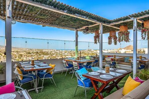 An outdoor dining area with tables and chairs, a view of the city, and a pergola with hanging baskets. - Cerro Colorado Condo | Condo in Cabo San Lucas (Cabo San Lucas)