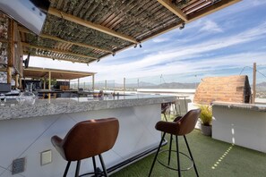 Eine Dachterrasse mit einer Steintheke, zwei Barhocker, einem Grill und Blick auf die Stadtlandschaft.