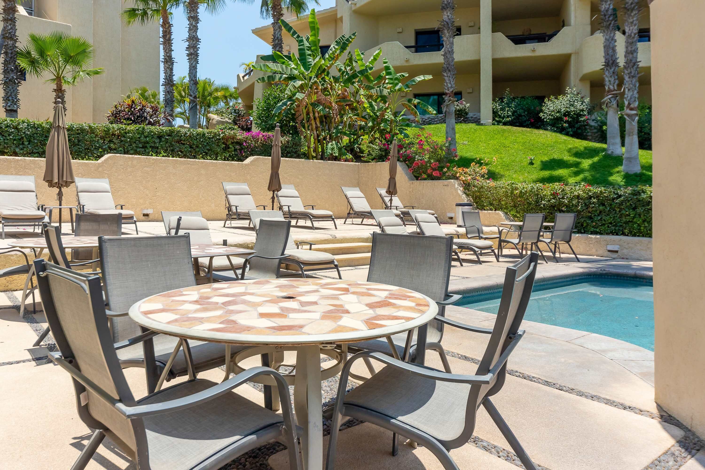 Un espace aménagé au bord de la piscine, avec des chaises longues et une table, entouré de palmiers et d’une pelouse bien entretenue.