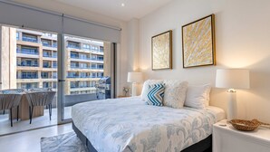 Habitación de hotel moderna con una cama grande, dos cuadros en la pared, un escritorio con sillas y un balcón con vistas a otros edificios.