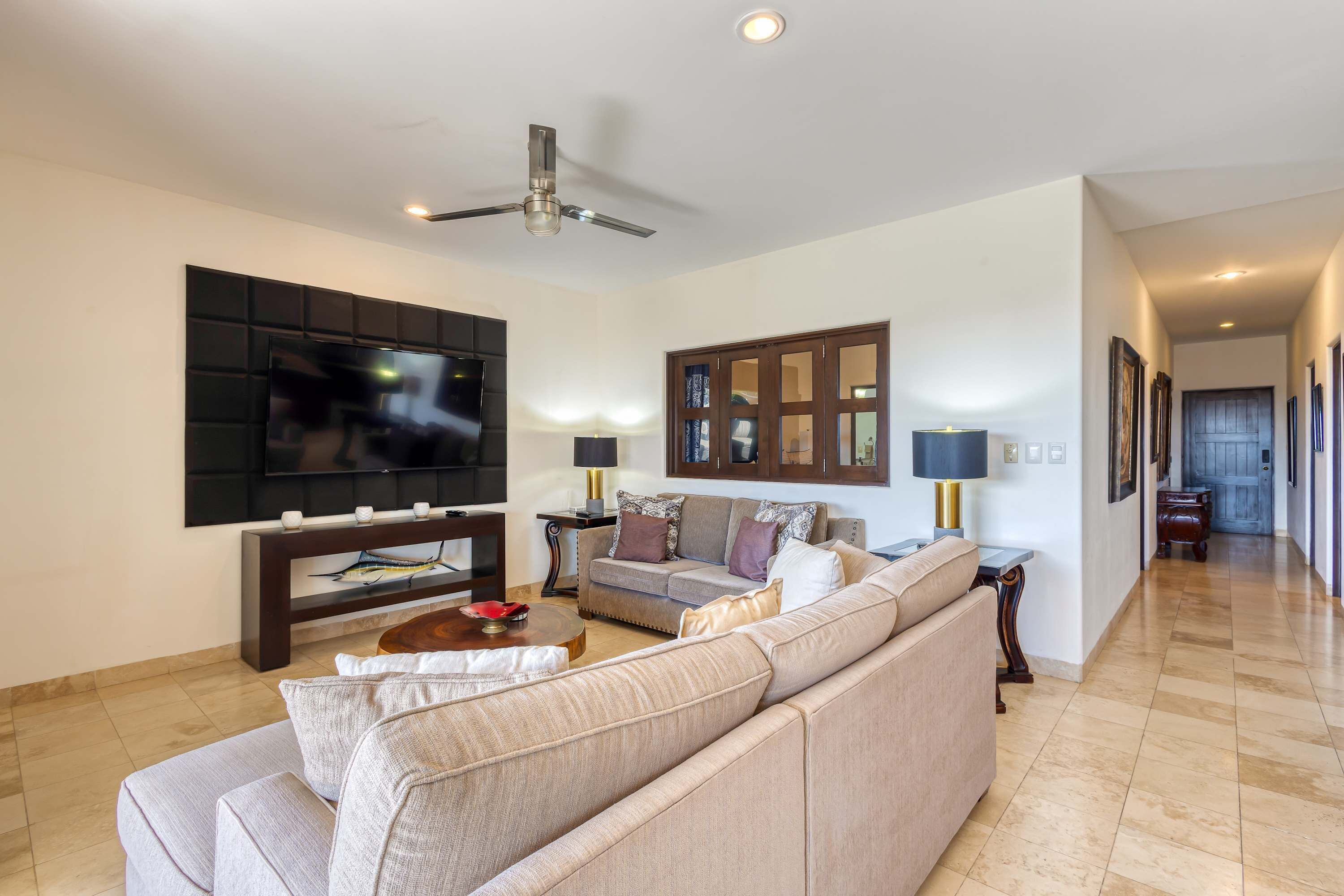 A living room with a flat-screen TV, a ceiling fan, a sofa, a coffee table, and a hallway with framed pictures.