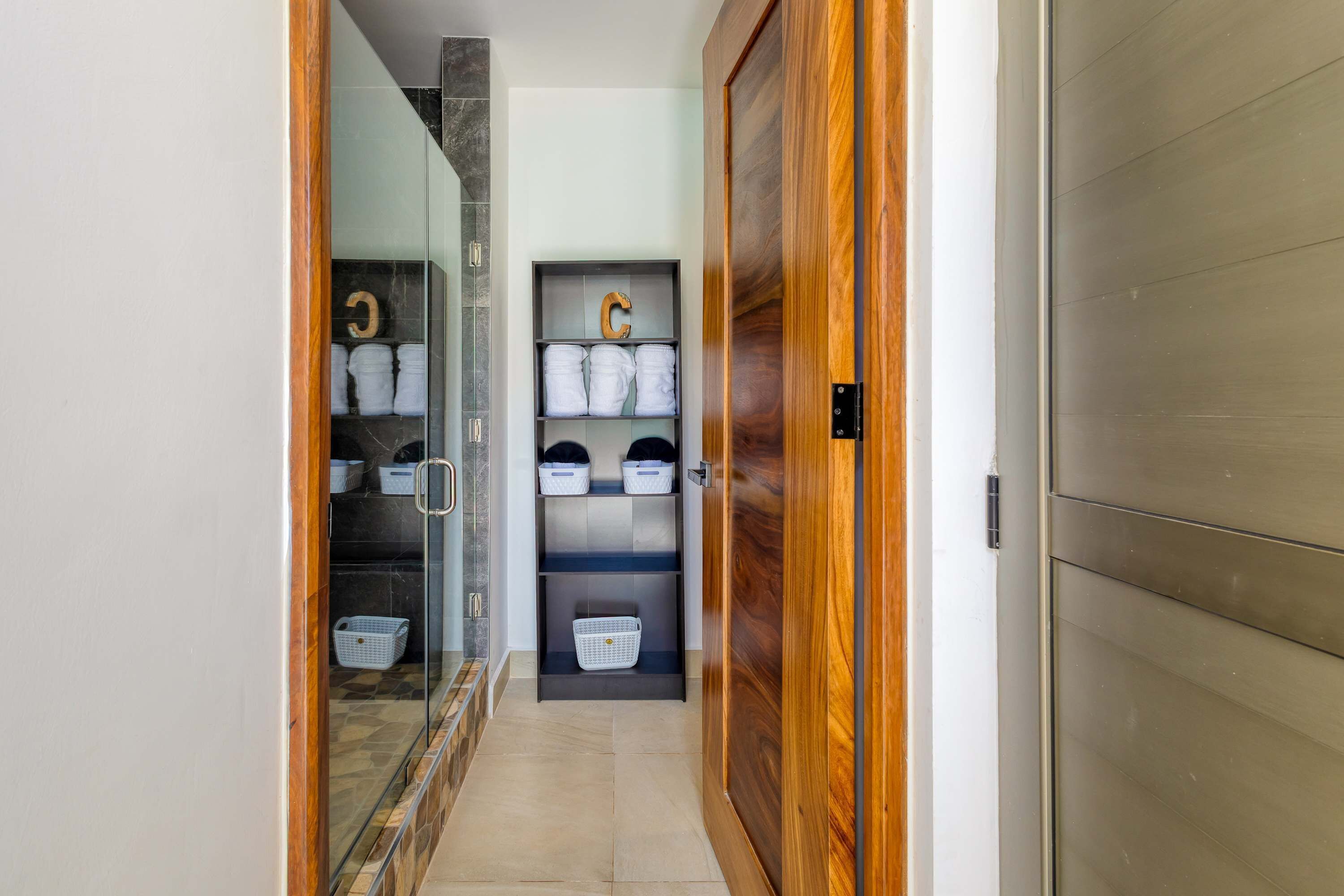 Een gang die leidt naar een kamer met een houten deur, een glazen douche en een opbergruimte met planken en manden.