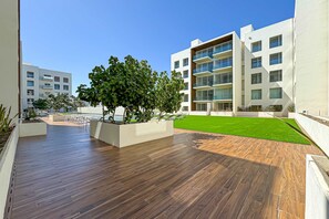 A modern outdoor area with a wooden deck, a grassy lawn, and a white building with balconies. - Luxury Condo | Gym, Rooftop Pool | Near Downtown (Cabo San Lucas)
