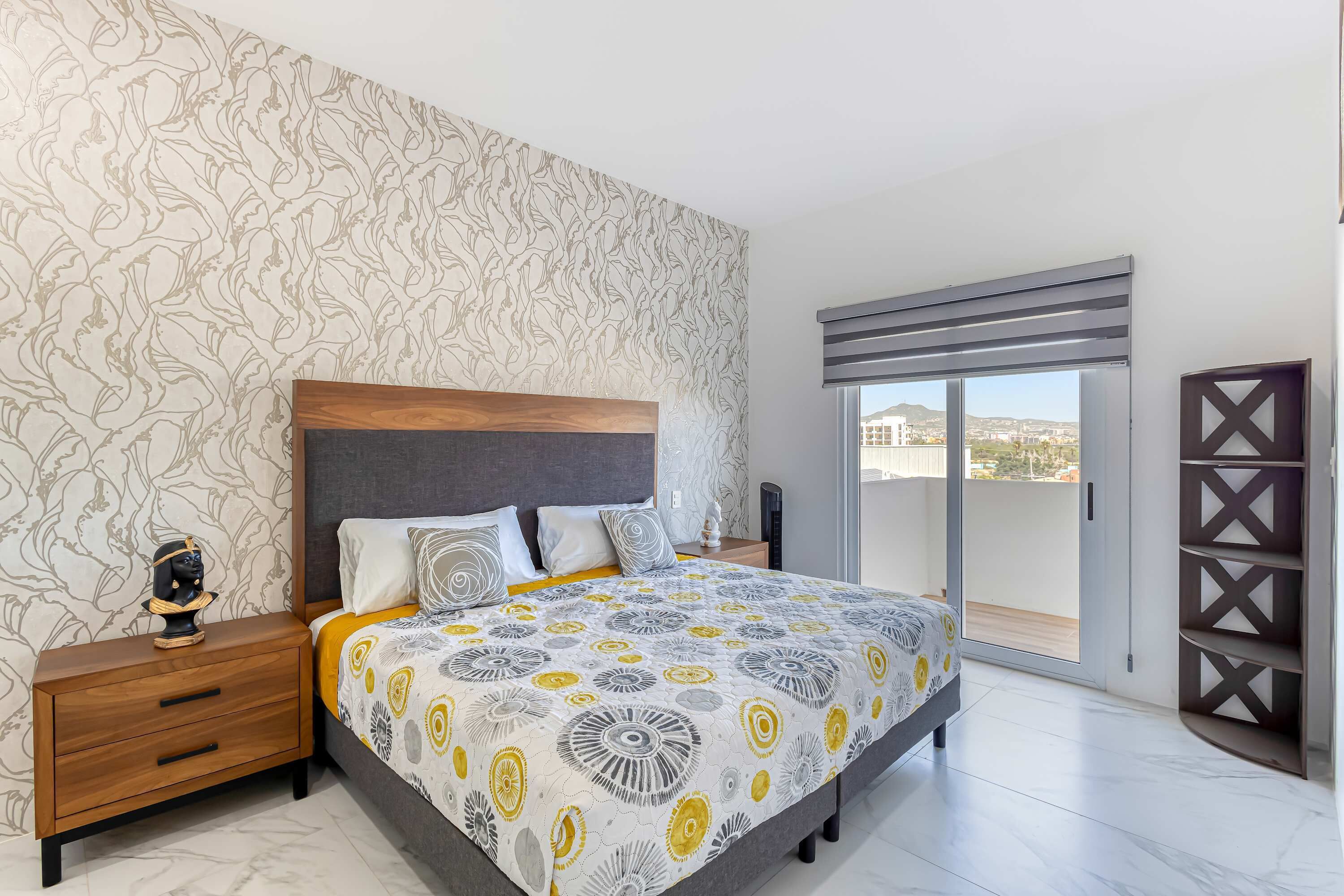 A bedroom with a patterned bedspread, a nightstand with drawers, a decorative item, and a view through a sliding door.