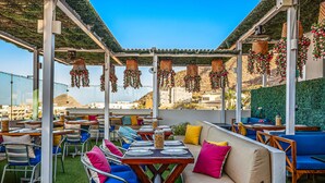 An outdoor dining area with tables and chairs, a sofa with cushions, hanging decorations, and a view of the landscape. - Morgan Residences 508A @ Boutique Hotel with pool, gym, spa (Cabo San Lucas)
