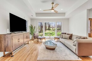 Ein modernes Wohnzimmer mit Flachbildfernseher, Holzschrank, Sofa, Couchtisch und Blick auf eine grüne Landschaft durch große Fenster.