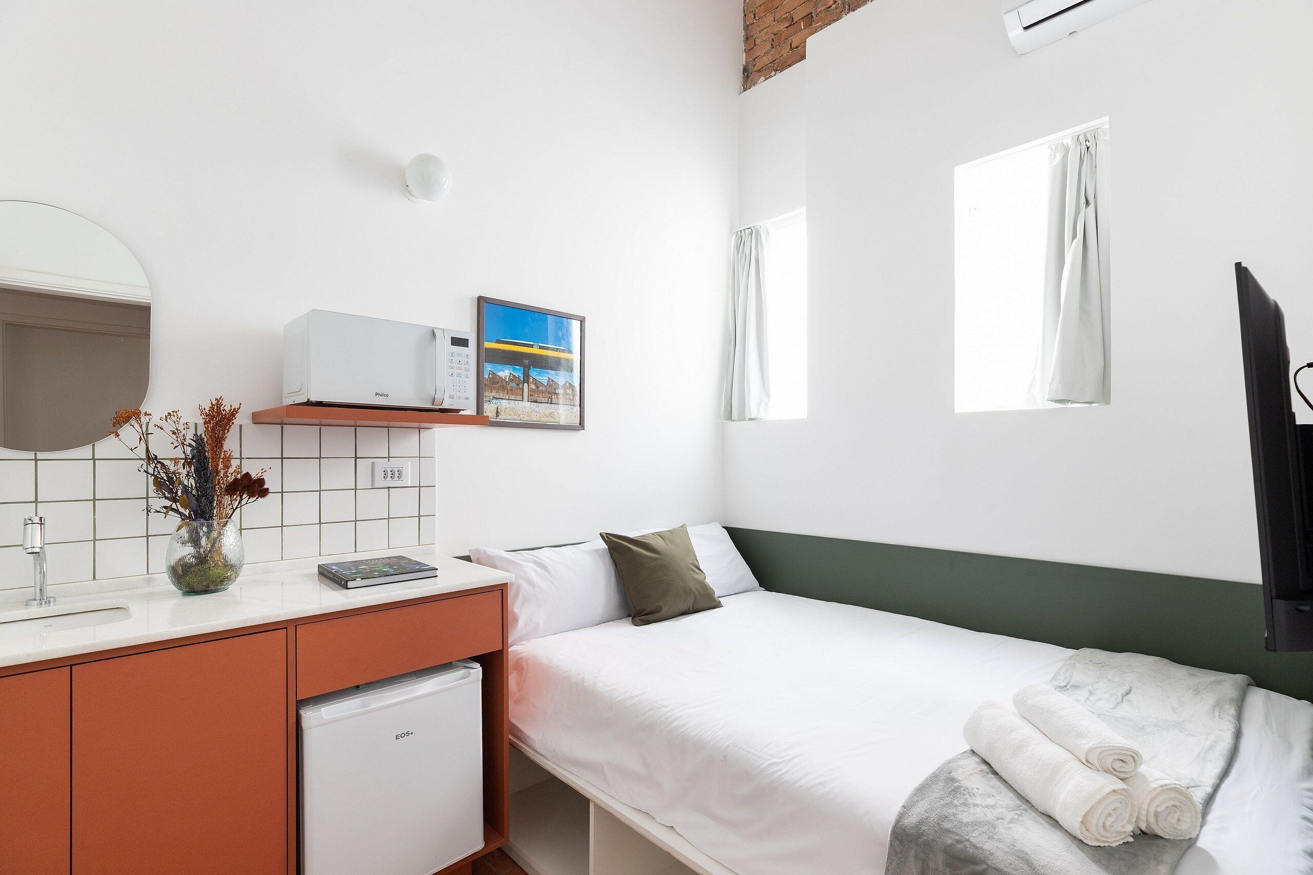 A hotel room with a bed, a small desk with a microwave, a sink, a mirror, and a window with curtains.