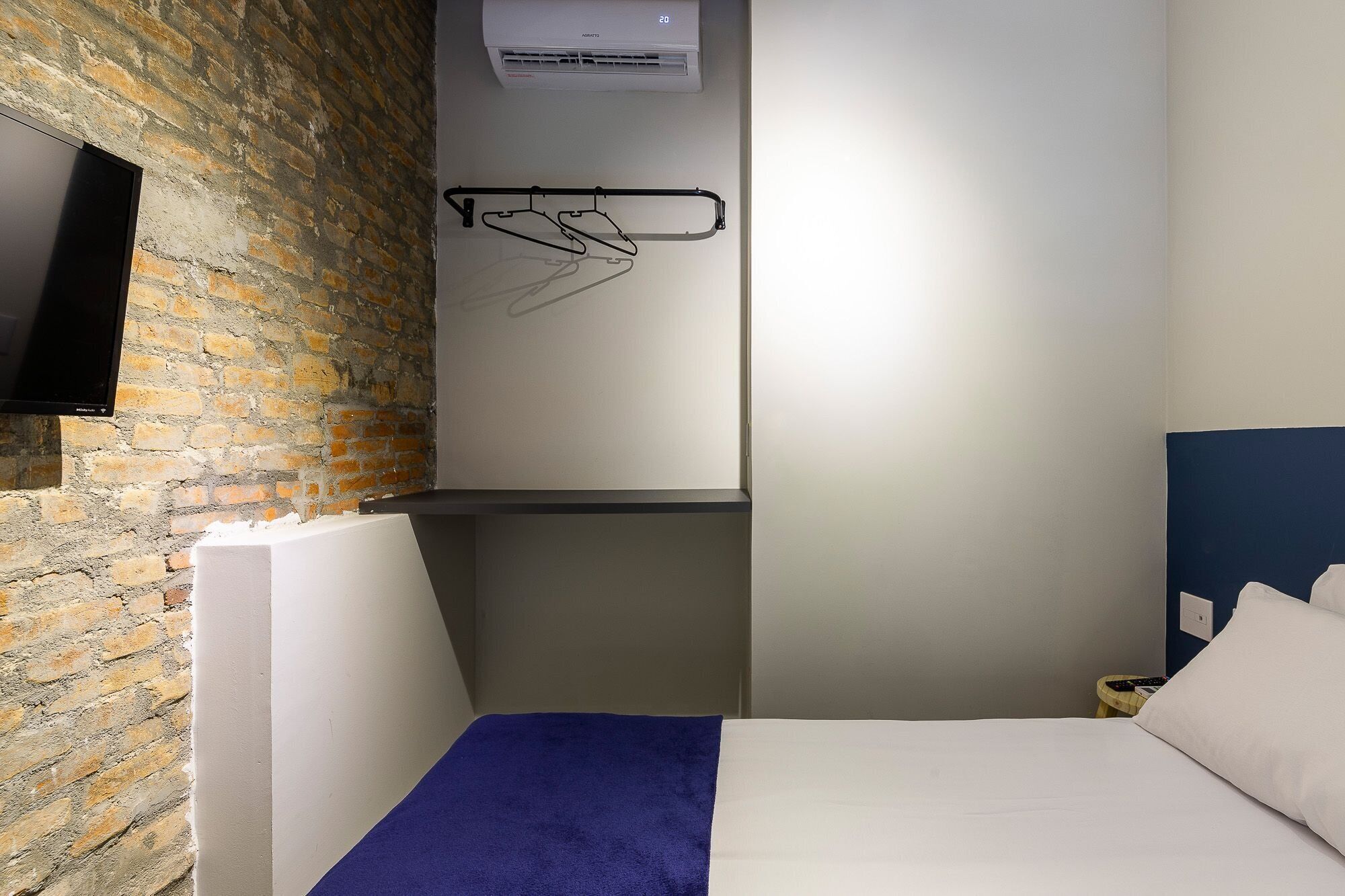 A hotel room with a brick wall, a mounted TV, a bed with a blue blanket, and a wall-mounted air conditioning unit.