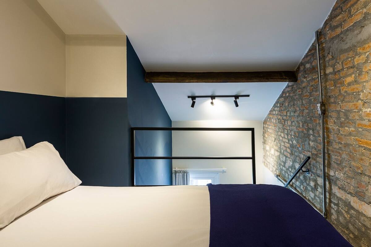 Un dormitorio con cama, cabecera, pared de ladrillo y techo con vigas a la vista.