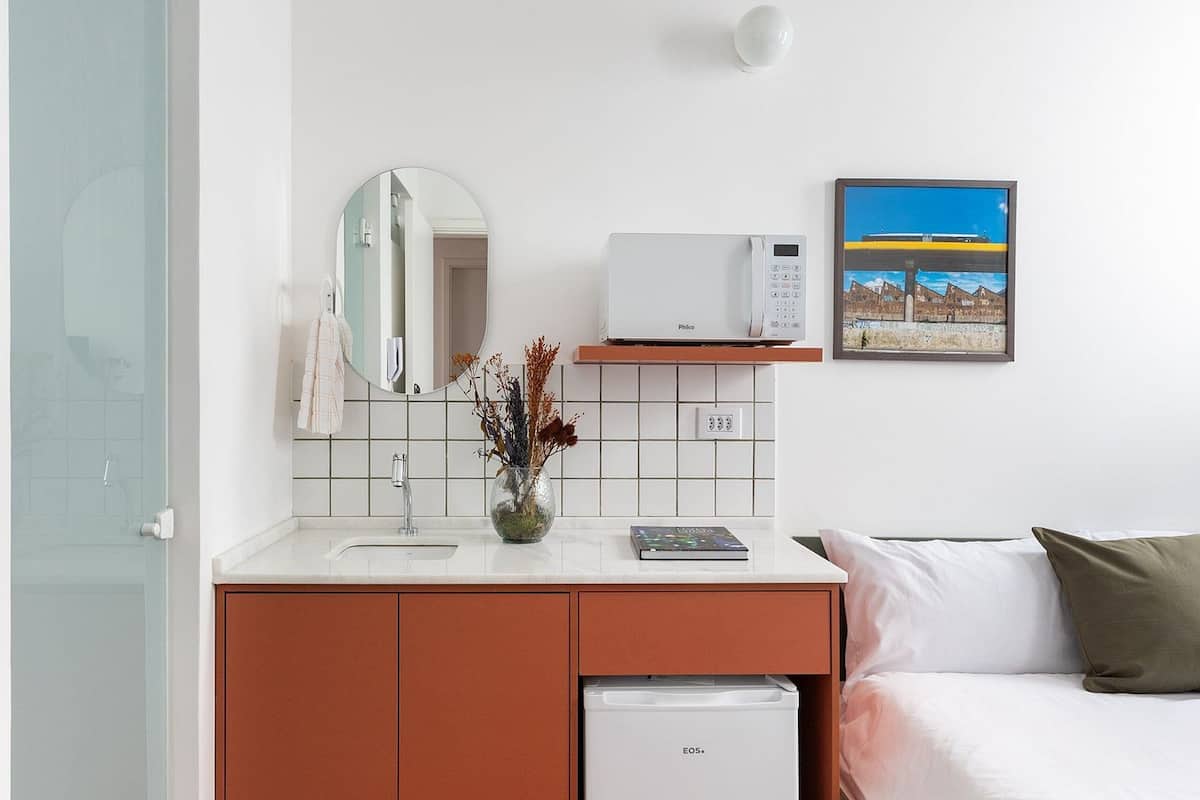 Habitación de hotel con un lavamanos blanco, microondas, refrigerador pequeño, cama con almohada verde y un cuadro en la pared.
