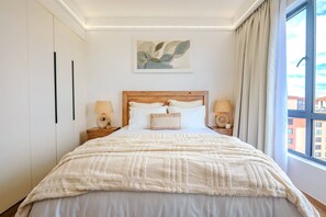 A bedroom with a bed, two bedside tables, a wall-mounted lamp, and a window with curtains.