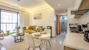 Sala de estar moderna com sofá, mesa de jantar e cadeiras, cozinha com fogão e utensílios, e uma janela grande.