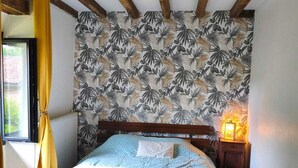 Un dormitorio con cama, vigas de madera, un papel tapiz estampado, una ventana con cortinas y una mesita de noche con lámpara.