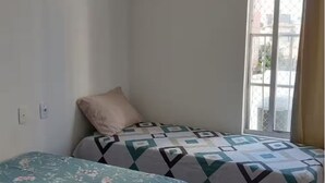 Una habitación pequeña con una cama individual, colcha estampada, una almohada, ventana con vista a edificios y un enchufe de pared.