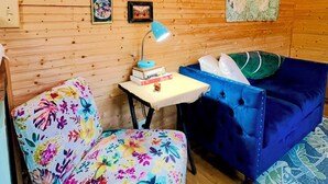 A cozy room with wooden walls, a blue sofa, a floral armchair, a small table with a lamp, and framed pictures on the wall. - Bamboo bungalow at Camp David. Pond-front, pet friendly tiny home retreat (Saluda)