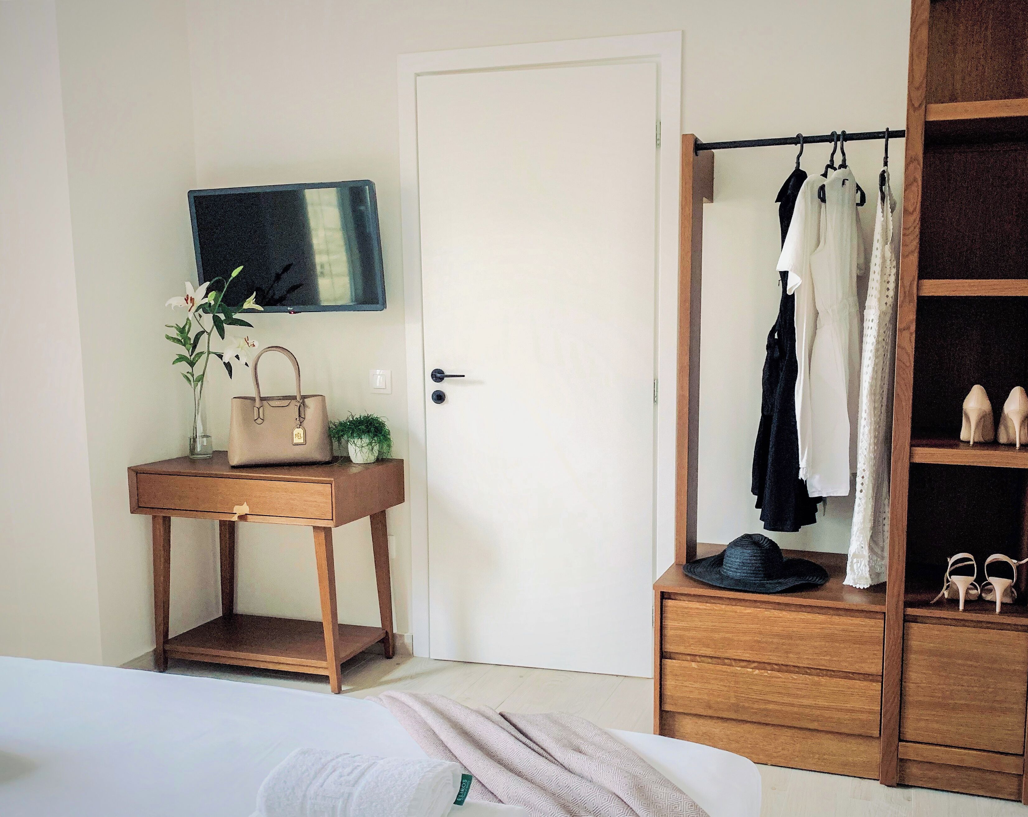Un dormitorio con cama, una mesita de noche de madera con un bolso y flores, un armario con ropa y zapatos, y una puerta con espejo.