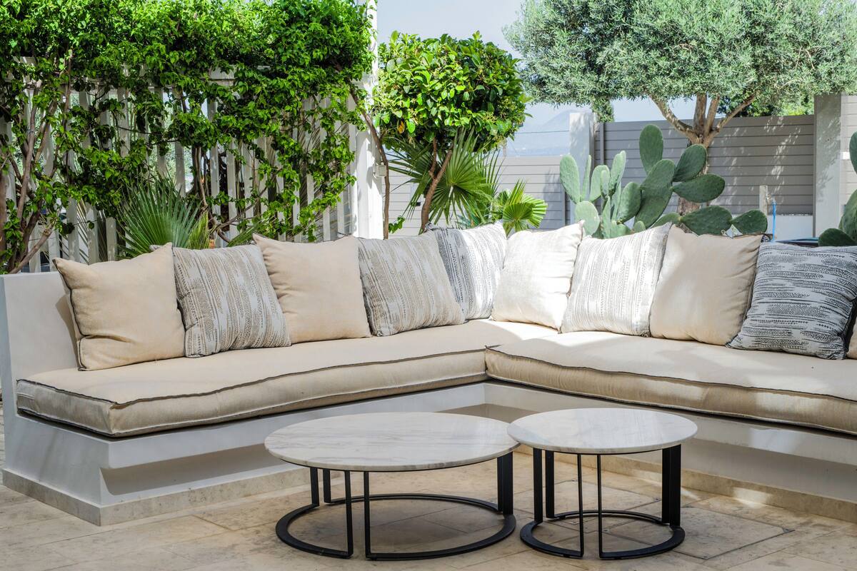 A sectional sofa with multiple pillows, two round tables, and a variety of plants.