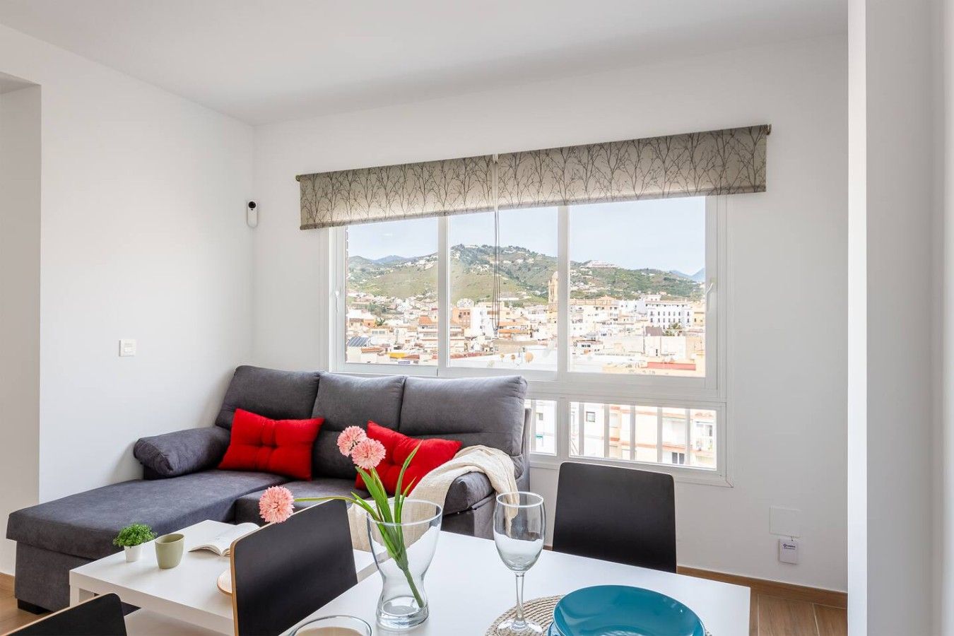 A modern living room with a grey sofa, red cushions, a small table with a vase of flowers, and a view of a cityscape through large windows.