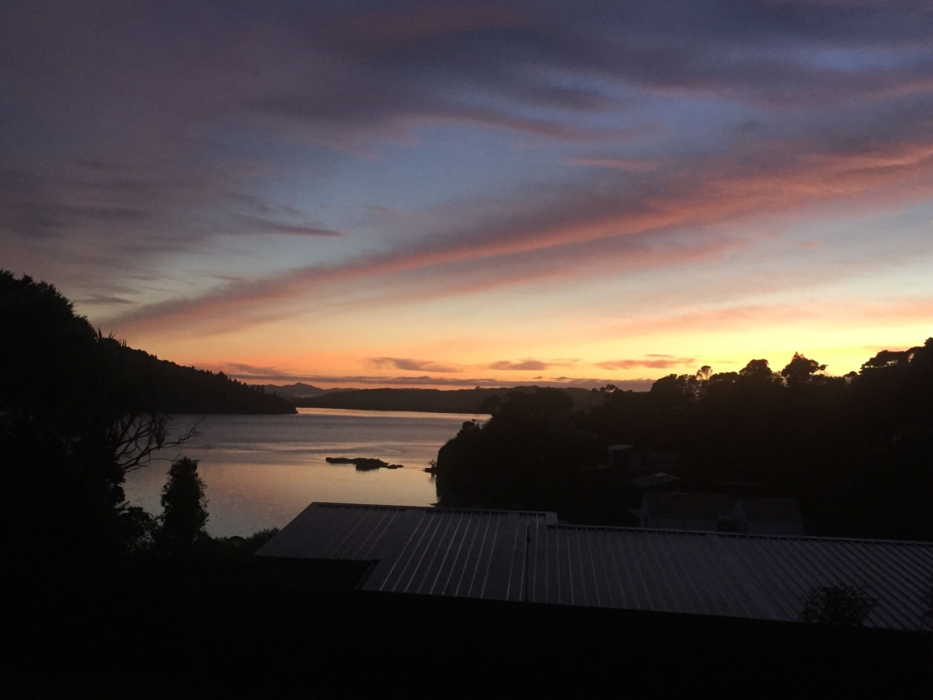 Un coucher de soleil sur un lac paisible, avec la silhouette d’une maison et d’arbres au premier plan.