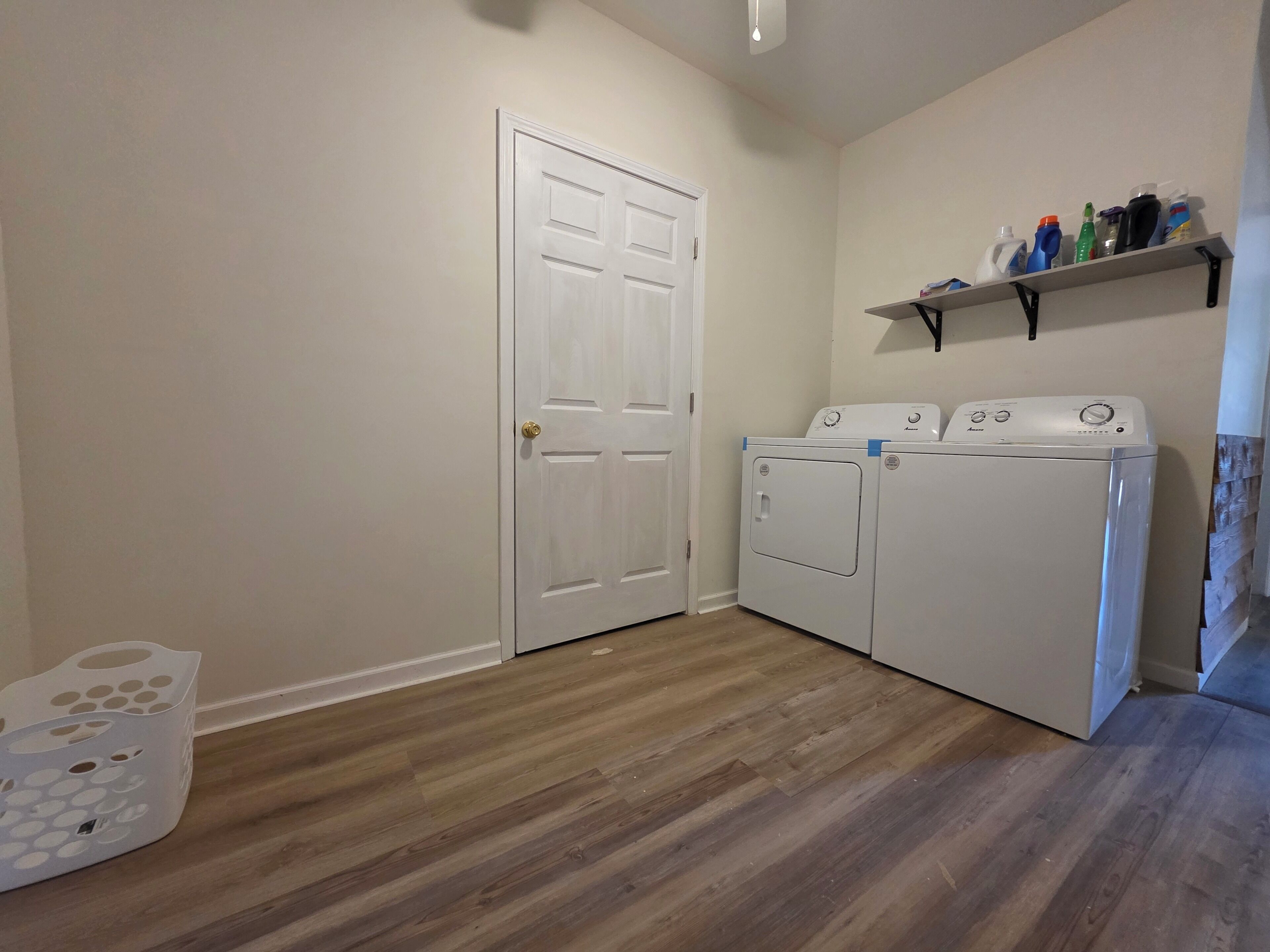 Une buanderie avec une porte blanche, deux machines à laver blanches, une étagère contenant des produits de nettoyage et un plancher en bois.