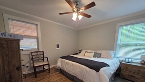 Quarto com cama, poltrona, cômoda, mesa de cabeceira, ventilador de teto e janela com persianas.