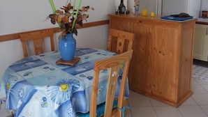 Un comedor con una mesa cubierta con mantel floreado, sillas de madera, un jarrón con flores, un aparador de madera, y una zona de cocina con un refrigerador y un espejo.