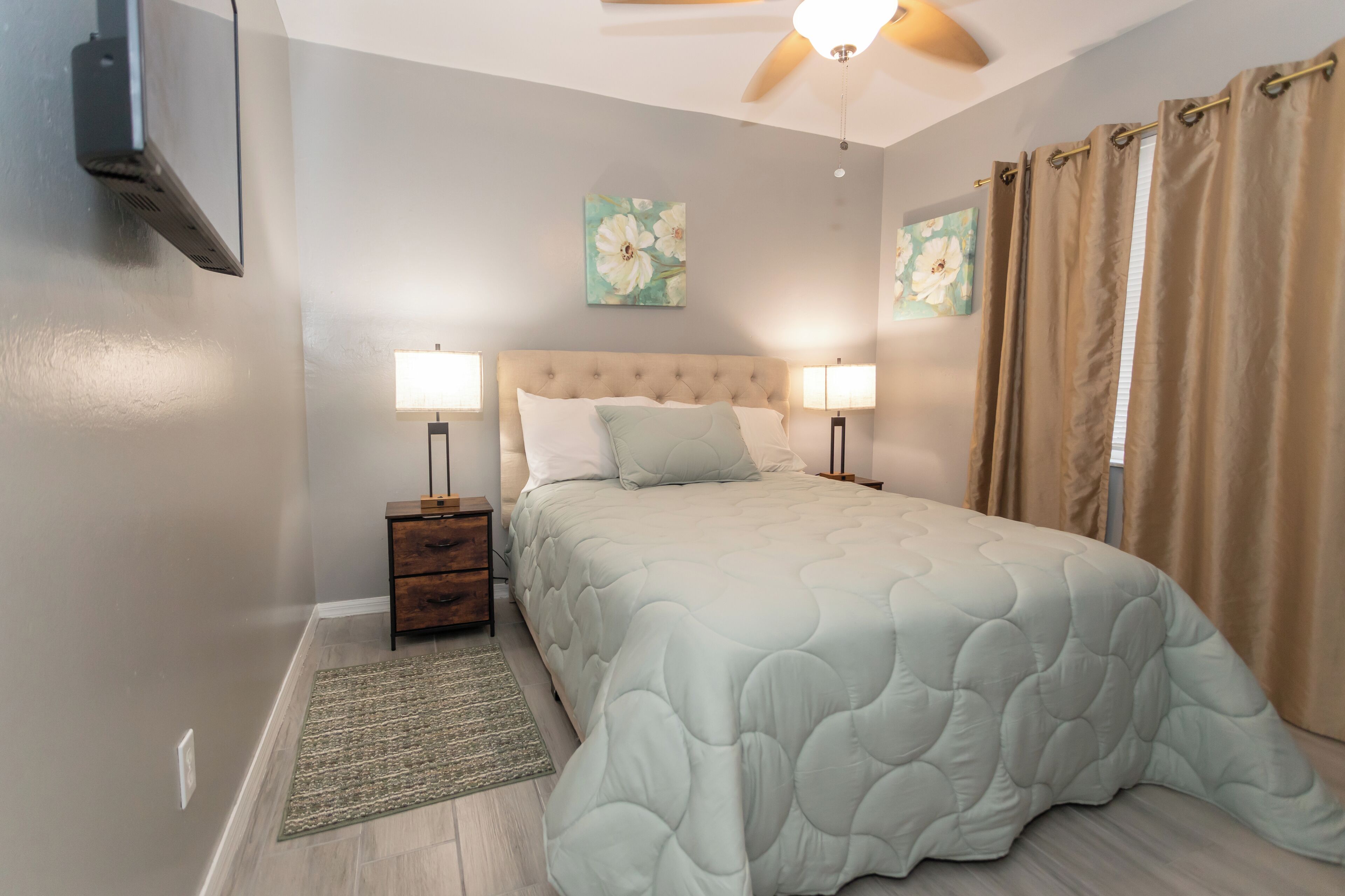 Un dormitorio con cama, lámparas de noche, mesitas de noche, ventilador de techo y dos cuadros colgados en la pared.
