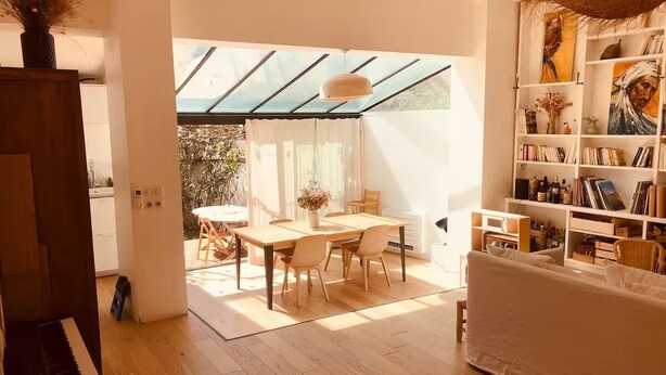 Ett modernt vardagsrum med ett piano, en matplats med bord och stolar samt en bokhylla fylld med böcker och dekorativa föremål.