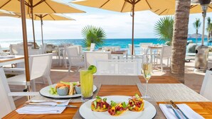 Ein Esstisch mit Tellern voller Essen, einem Glas Weißwein und einem Cocktail mit Limettenscheibe, vor einer Strand- und Meerlandschaft.