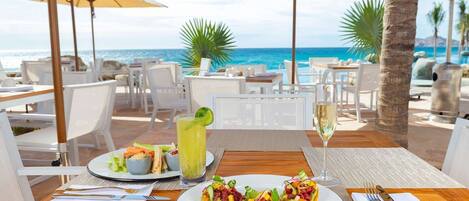 A dining table with plates of food, a glass of white wine, and a cocktail with a lime garnish, set against a backdrop of a beach and ocean view.