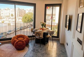 Comedor con mesa dispuesta para cuatro, ventanas amplias con vista a la ciudad, una silla roja y un televisor de pantalla plana montado en la pared.