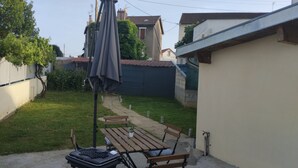 A patio with a grill, wooden table, and chairs, surrounded by a garden and a blue garage door. - Iglu Beautiful apartment, two comfortable bedrooms, outdoor camera. Garden. (Villeneuve-le-Roi)