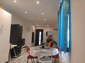 Un coin repas avec une table ronde, des chaises blanches, un réfrigérateur noir et un rideau bleu.