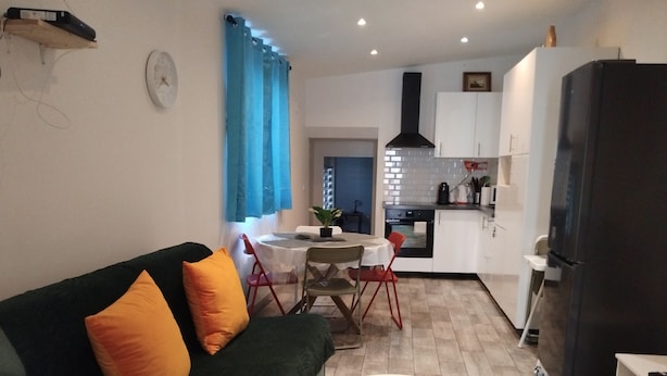 A living room with a sofa, a dining table with chairs, a kitchen area with cabinets and appliances, and a small table with a plant and a book. - Iglu Beautiful apartment, two comfortable bedrooms, outdoor camera. Garden. (Villeneuve-le-Roi)