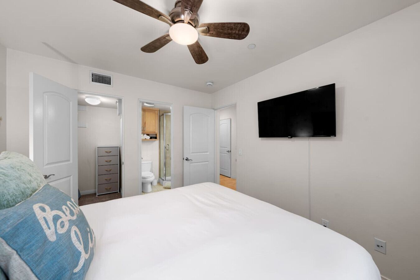 Een slaapkamer met een bed, een plafondventilator, een televisie en een zichtbare badkamer door een geopende deur.